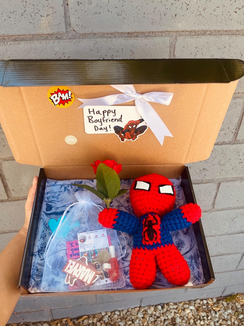 May include: A brown cardboard box with a white ribbon and a "Happy Boyfriend Day!" card featuring a cartoon Spiderman. The box is open and contains a red and blue crocheted Spiderman doll, a red rose, a small bag with a "Marvel" sticker, and a small pink card.