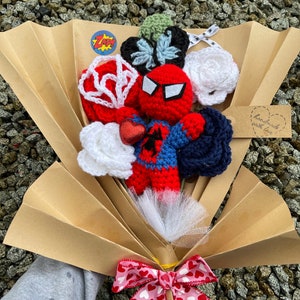 May include: A crocheted bouquet featuring a Spiderman character, white roses, a blue and white butterfly, and a red and white web. The bouquet is wrapped in brown paper and tied with a pink ribbon with white hearts. The tag says "handmade with love".
