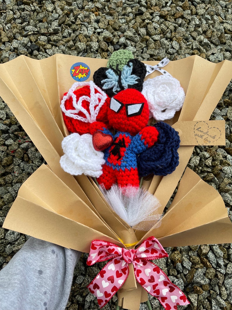 May include: A crocheted bouquet featuring a Spiderman character, white roses, a blue and white butterfly, and a red and white web. The bouquet is wrapped in brown paper and tied with a pink ribbon with white hearts. The tag says "handmade with love".
