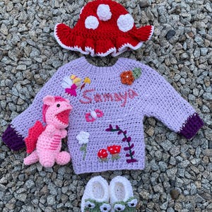 Può includere: Un set di abbigliamento per neonati all'uncinetto fatto a mano, tra cui un cappello a fungo rosso con pois bianchi, un maglione lavanda con il nome "Samaya", un drago rosa e scarpine per neonati a tema rana. Il maglione ha abbellimenti floreali e a fungo.