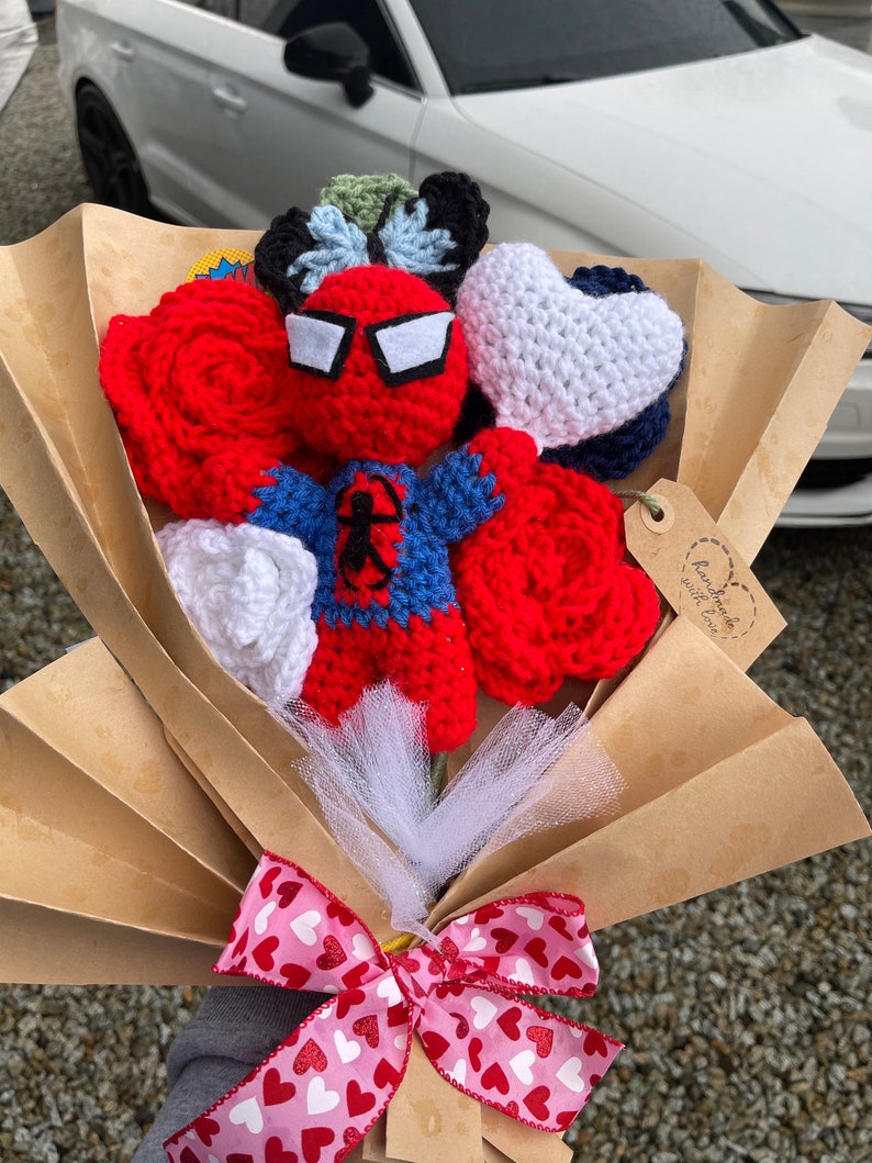 May include: A crocheted bouquet featuring a red and blue Spiderman character, red roses, white hearts, and a blue and white heart. The bouquet is wrapped in brown paper and tied with a pink ribbon with white hearts.