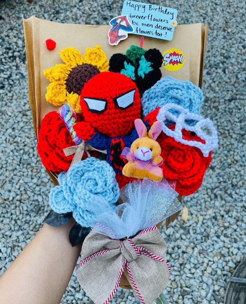 May include: A crocheted bouquet of flowers with a red crocheted Spiderman figure in the center. The bouquet also includes a crocheted bunny, a crocheted pair of glasses, and a crocheted sunflower. The bouquet is wrapped in brown paper and tied with a brown ribbon. The bouquet is sitting on a table with a note that says "Happy Birthday forever flowers bc men deserve flowers too!"
