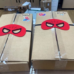 May include: Two brown cardboard boxes with red felt Spider-Man masks taped to the top. The boxes are labeled "Cameron" and "Wendy".