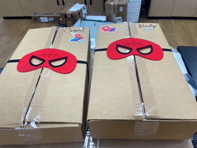 May include: Two brown cardboard boxes with red felt Spider-Man masks taped to the top. The boxes are labeled "Cameron" and "Wendy".