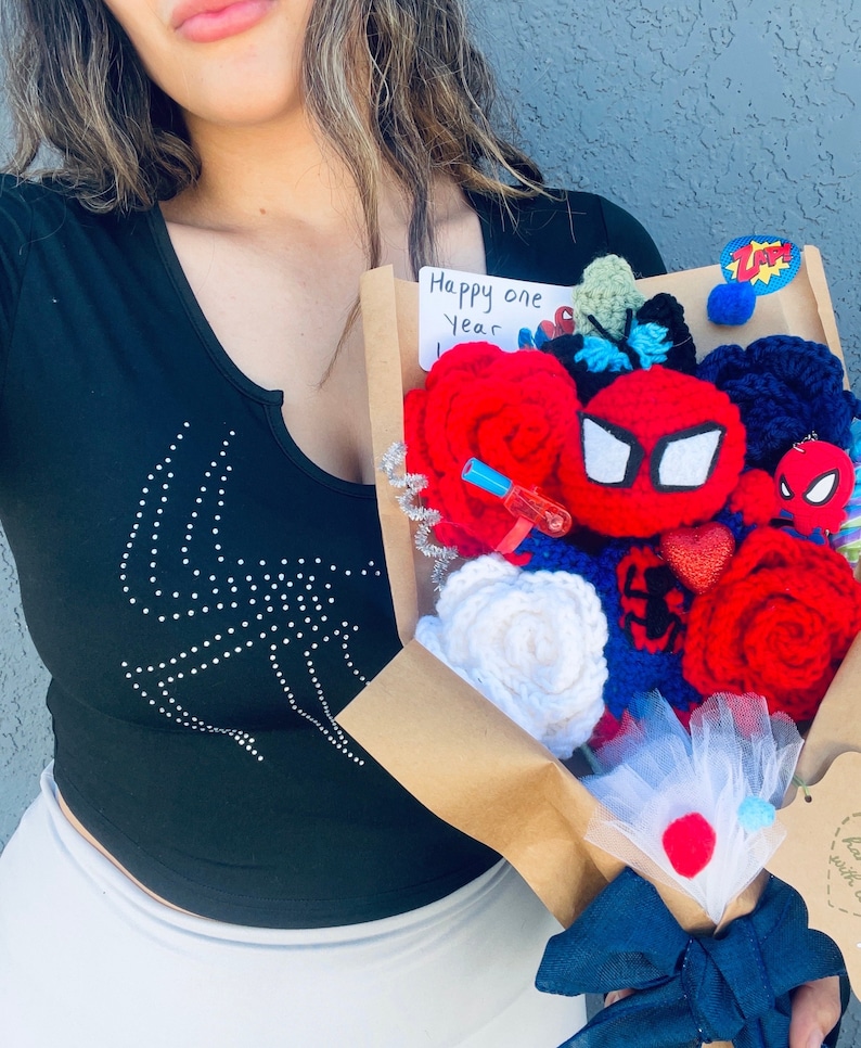 May include: A brown paper bouquet with a red, white, and blue crocheted flower arrangement. The bouquet includes a red crocheted Spider-Man mask, a red crocheted Spider-Man figure, and a small red crocheted heart. The bouquet also includes a small white card with the text "Happy one year".