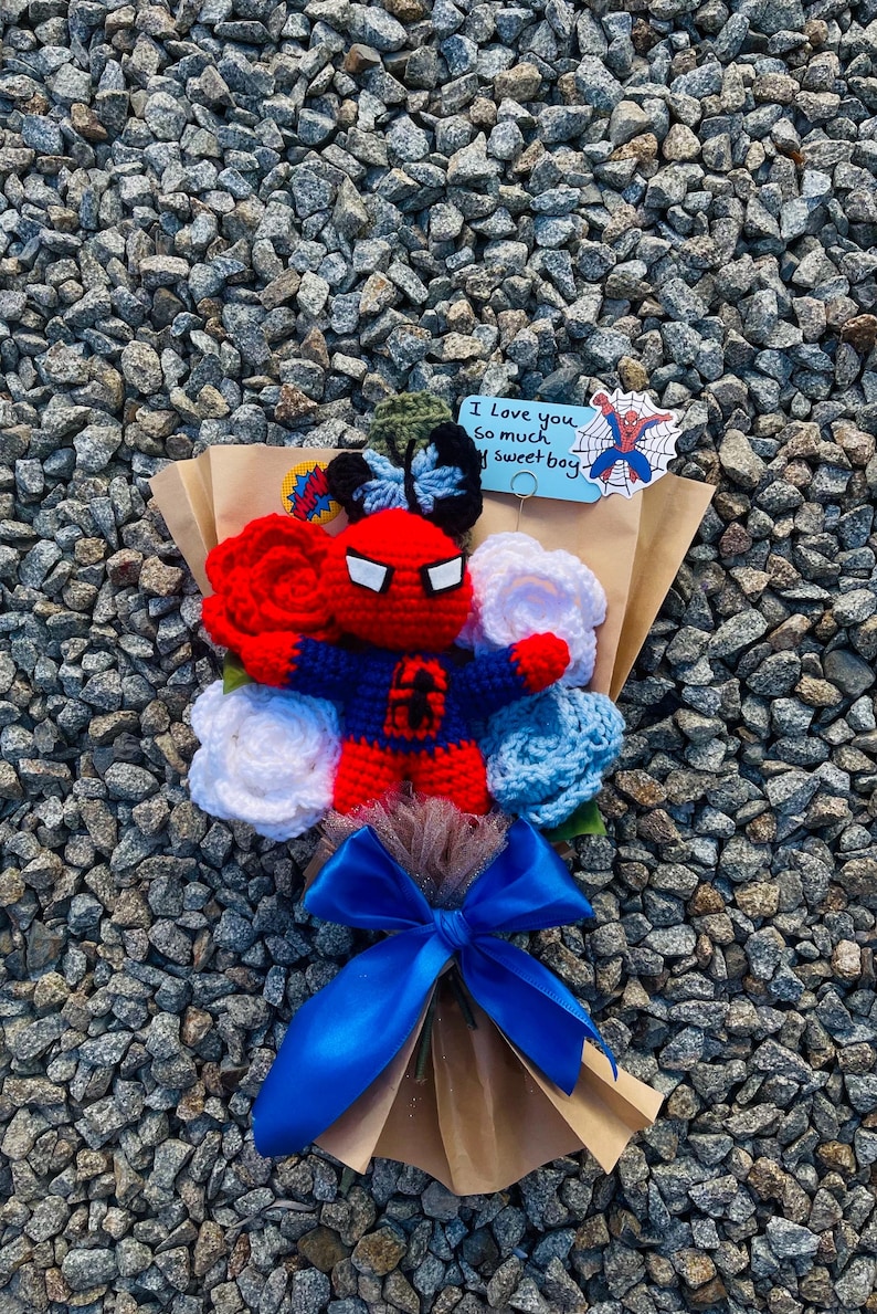 May include: A crocheted bouquet featuring a red and blue Spiderman character, white and blue crocheted flowers, and a blue ribbon. The bouquet is wrapped in brown paper and includes a card that says "I love you so much my sweet boy".