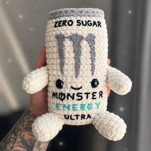 Può includere: Un peluche all'uncinetto bianco a forma di lattina con la parte superiore grigia. Presenta il testo ricamato ZERO SUGAR, MONSTER ENERGY e ULTRA, un logo a forma di artiglio grigio, occhi di sicurezza neri e un sorriso cucito. Ha due piccole braccia e gambe.