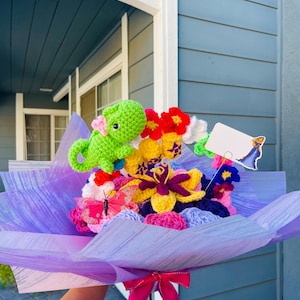 May include: A crocheted bouquet of flowers, a butterfly, and a green dinosaur. The bouquet is wrapped in purple tissue paper and tied with a pink ribbon.