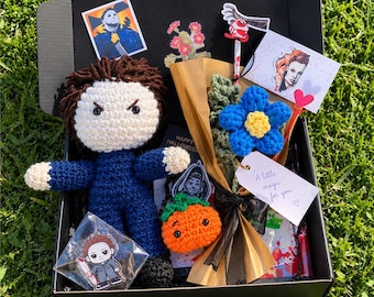May include: A black gift box containing a crocheted doll with brown hair and a blue jumpsuit, a small crocheted orange pumpkin, a crocheted blue flower bouquet wrapped in brown paper, stickers, a card with text A little magic for you, and a pin.