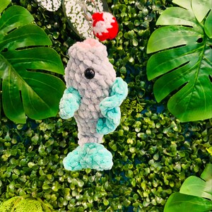 May include: A crocheted blue and gray fish keychain with a red pom-pom on top. The keychain is hanging from a silver key ring.
