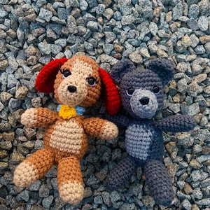 May include: Two crocheted stuffed animals, a brown and tan dog with red ears and a gray bear, are lying on a bed of gray gravel.