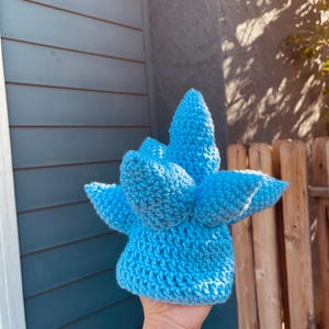 May include: A handmade, light blue crochet hat with a unique, spiky design. The hat is held up against a blue and wooden background, showcasing its textured, three-dimensional form. The hat's color is a vibrant, solid blue.