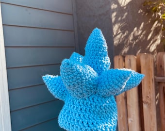 May include: A handmade, light blue crochet hat with a unique, spiky design. The hat is held up against a blue and wooden background, showcasing its textured, three-dimensional form. The hat's color is a vibrant, solid blue.