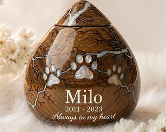 May include: A teardrop-shaped pet urn made of polished brown wood with a natural grain and white lightning-like resin veins. It features three inlaid mother-of-pearl paw prints and engraved text reading Milo, 2011-2023, and Always in my heart.