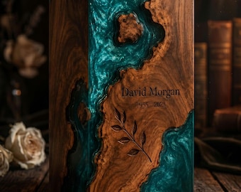 May include: A rectangular wooden cremation urn featuring a swirling, marbled teal resin inlay that mimics a river flow. The wood surface is engraved with the name David Morgan, the dates 1955-2023, and a delicate, detailed botanical leaf branch.