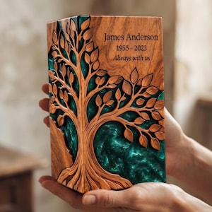 May include: A rectangular wooden memorial urn featuring a carved tree design with intricate branches and leaves. The wood grain is visible, and the background of the tree is filled with a swirling teal resin. The text reads James Anderson 1955-2023 Always with us.