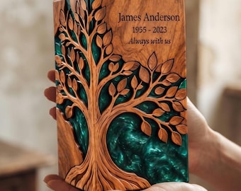 May include: A rectangular wooden memorial urn featuring a carved tree design with intricate branches and leaves. The wood grain is visible, and the background of the tree is filled with a swirling teal resin. The text reads James Anderson 1955-2023 Always with us.