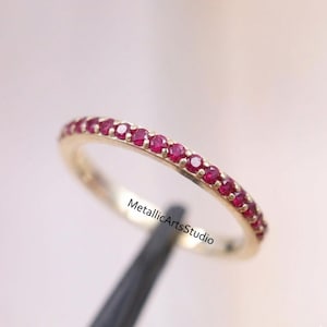 Può includere: Un anello a fascia dorata con piccole gemme rosse vibranti. L'anello è sottile e delicato, con le gemme che circondano la fascia. Il testo "MetallicArtsStudio" è visibile sull'anello.