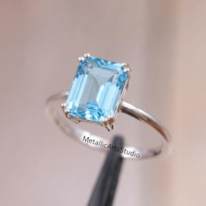 Pode incluir: Um anel de prata com uma grande pedra preciosa retangular azul clara. A pedra preciosa é definida em uma configuração de quatro pontas, e a banda é de prata polida simples. O anel é exibido contra um fundo macio e desfocado.