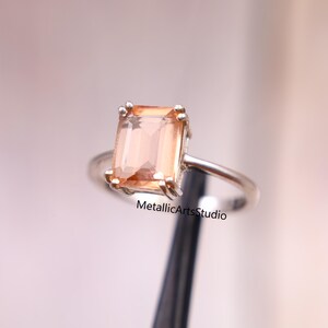 Puede incluir: Un anillo de plata con una piedra preciosa rectangular de color melocotón. La piedra está engastada en un engaste de cuatro garras. El anillo es sencillo y elegante, con un acabado pulido. El texto "MetallicArtsStudio" es visible.