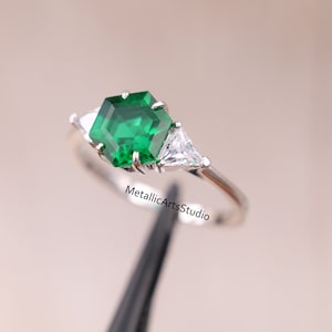 Puede incluir: Un anillo de plata con una esmeralda verde hexagonal y dos piedras preciosas triangulares transparentes más pequeñas.  MetallicArtsStudio.