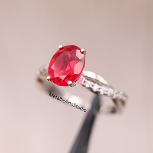 Könnte beinhalten: Ein silberner Ring mit einem großen, oval geschliffenen Rubin als Mittelstein. Das Band hat ein verdrehtes Design mit kleinen, klaren Akzentsteinen. Der Ring ist vor einem weichen, neutralen Hintergrund platziert.