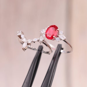 May include: A silver ring with a large red oval gemstone surrounded by smaller clear gemstones. The ring is set on a black background.