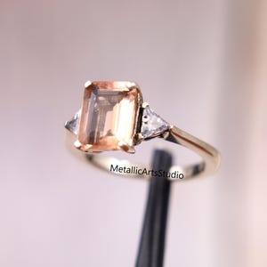 Puede incluir: Un anillo de oro con una piedra preciosa rectangular de color melocotón como punto focal principal. Dos piedras preciosas triangulares y transparentes más pequeñas flanquean la piedra principal. El anillo tiene una banda de oro pulido. El texto "MetallicArtsStudio" es visible.