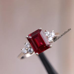Puede incluir: Un anillo de oro con una gran gema rectangular de color rojo intenso. Gemas más pequeñas y claras se agrupan alrededor de la piedra principal. El anillo está sobre un fondo borroso.