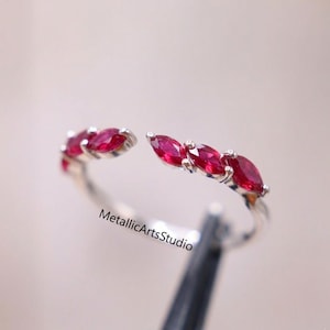 May include: Open-ended silver ring featuring five marquise-cut, vibrant red gemstones. The ring's design is modern and elegant, with the gemstones set in a row across the top. The ring is a piece of jewelry.