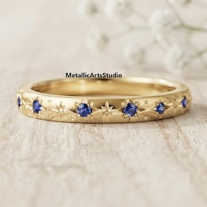 May include: A gold band ring featuring a series of round blue sapphire gemstones alternating with engraved starburst patterns. The metal surface is polished and smooth, with the text MetallicArtsStudio visible above the ring.