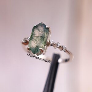 Puede incluir: Anillo de plata con una piedra de ágata musgosa rectangular, con patrones verdes y blancos. La banda está adornada con pequeñas piedras claras. El texto "MetallicArtsStudio" es visible en el anillo.