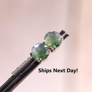May include: A pair of round, green moss agate stud earrings with a silver setting. The earrings are displayed on a black surface. The text "MetallicArtsStudio" and "Ships Next Day!" are visible.