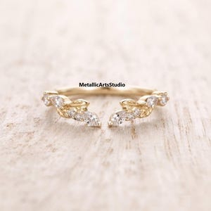 May include: A gold open ring featuring a leaf-inspired design with small clear stones set along the band. The ring has a gap in the center and displays the text MetallicArtsStudio above the band. The metal has a polished yellow gold finish.