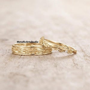 Puede incluir: Dos anillos de oro con un intrincado patrón de hojas botánicas en relieve en toda la superficie. La banda más ancha está colocada horizontalmente, mientras que la banda más fina descansa en ángulo, mostrando una pequeña piedra preciosa redonda y clara.