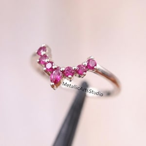May include: A silver ring with a curved design, set with multiple vibrant pink gemstones. The ring's band is thin, and the gemstones are arranged in a delicate, arching pattern. The ring is a piece of jewelry.