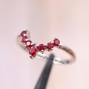 Peut inclure: Une bague en argent avec un design incurvé, ornée de plusieurs pierres précieuses rouges ovales et rondes. La bague porte le texte "MetallicArtsStudio". Les pierres sont serties de près, créant un effet scintillant continu.