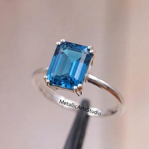 May include: A silver ring featuring a large, rectangular, vibrant blue gemstone. The gemstone is the focal point, set in a classic four-prong setting. The ring band is a simple, polished silver. The text "MetallicArtsStudio" is visible.