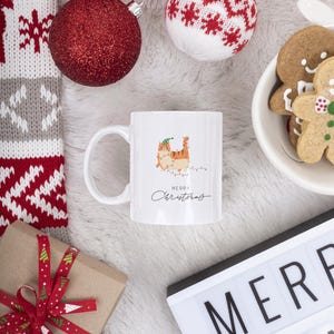 Puede incluir: Taza de cerámica blanca con una ilustración de gato y el texto "Merry Christmas". La taza está rodeada de adornos navideños, un regalo envuelto con una cinta roja y galletas de jengibre.
