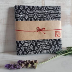 Può includere: Un pacco avvolto con un tessuto a motivi geometrici bianchi e neri. È assicurato con una fascia di carta marrone e spago rosso legato con un fiocco. Un rametto di lavanda è vicino. Il pacco ha un timbro giapponese.