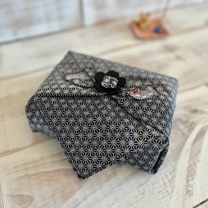May include: A folded fabric item with a black and white geometric pattern. A black flower-shaped embellishment is attached to the top. The item is resting on a light-colored wooden surface.
