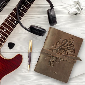 May include: A brown leather journal with a music note design and the word "notes" embossed on the cover. A guitar, headphones, and a pen are also in the image.