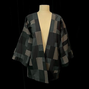 May include: A patchwork jacket in shades of black and gray, displayed on a mannequin. The jacket features a geometric pattern of rectangular shapes, with a loose, open front design and wide sleeves. The fabric appears to be a soft, woven material.