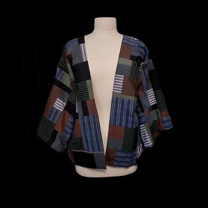 May include: A patchwork jacket with a variety of colors and patterns, including brown, blue, green, and black. The jacket is displayed on a mannequin against a black background. The jacket has an open front and three-quarter sleeves.