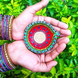 Puede incluir: Una diya colorida, una lámpara de aceite tradicional india, con pintura verde, azul, roja y dorada. La diya está siendo sostenida en las palmas de las manos de una persona.
