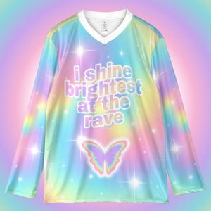 May include: A long-sleeved, V-neck shirt with a vibrant, rainbow-colored design. The shirt features the text "i shine brightest at the rave" and a butterfly graphic. The design includes sparkling star accents.
