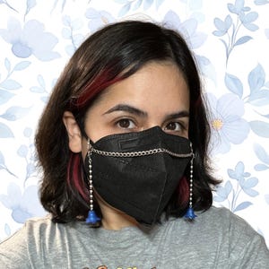 Könnte beinhalten: Schwarze Gesichtsmaske mit einer silbernen Kette und baumelnden blauen Blumenanhängern. Die Maske wird mit einem grauen T-Shirt getragen. Der Hintergrund zeigt ein Blumenmuster mit blauen Blumen.