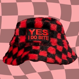 Peut inclure: Chapeau bob en fourrure rouge et noir à carreaux avec un patch noir qui dit "YES I DO BITE" en lettres rouges.