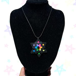 Puede incluir: Un collar con colgante negro en forma de estrella. El colgante está hecho de pequeñas cuentas de colores y cuelga de una cadena plateada. El collar se exhibe en un maniquí negro.