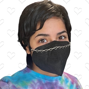 May include: A person wearing a black face mask with a silver heart-shaped chain attached to the ear loops. The person is wearing a tie-dye shirt.